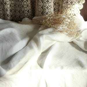 May include: A still life composition featuring a white pillar candle, delicate dried flowers, and a flowing white fabric. The fabric is draped over a surface, creating soft folds and shadows. A lace curtain is visible in the background.