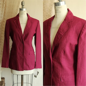 May include: A deep magenta blazer featuring a notched lapel, long sleeves, and a two-button front closure. The garment includes two front flap pockets and is displayed on a mannequin, showcasing a textured fabric finish and a tailored fit.