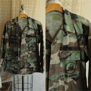May include: A long-sleeve camouflage jacket featuring a woodland pattern in shades of green, brown, and black. The jacket includes a collar, button-down front, and four front pockets. A circular patch with a bee graphic and the text SEABEES is present.