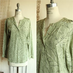 Könnte beinhalten: Eine hellgrüne Langarmbluse mit V-Ausschnitt und Knopfverschluss. Die Bluse hat ein Muster aus weißen und schwarzen blattartigen Designs. Das Kleidungsstück wird auf einer Schaufensterpuppe präsentiert.