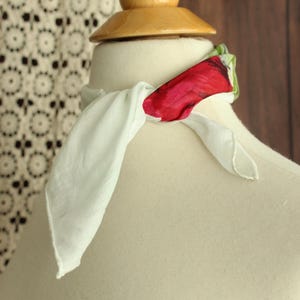 以下が含まれることがあります： マネキンの首に巻かれた白いスカーフ。鮮やかな赤と緑の花柄が特徴です。スカーフは軽量の生地でできており、端はゆるく垂れ下がっています。マネキンは木製の首と頭をしています。