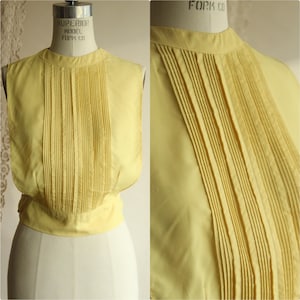 Puede incluir: Top sin mangas amarillo pálido con pliegues verticales en la parte delantera. El top tiene un cuello alto y redondeado y una cintura entallada. La tela parece ser un material ligero. El top se muestra en un maniquí.