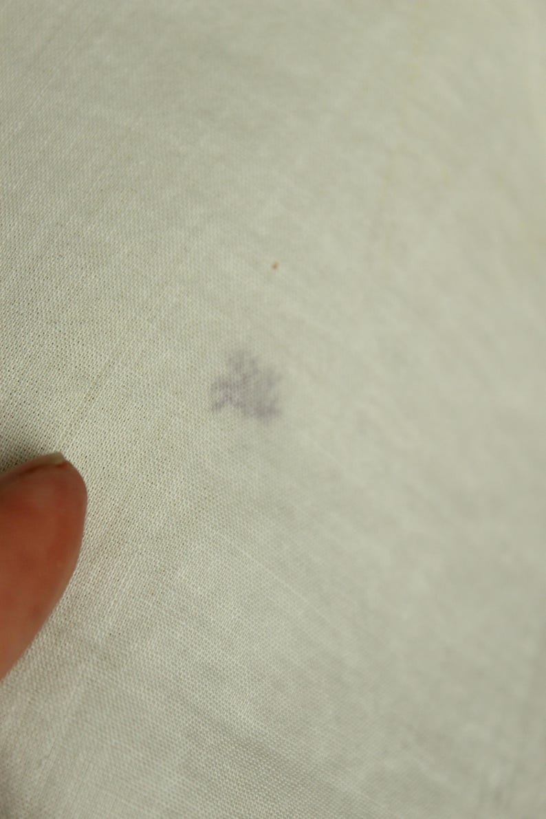 以下が含まれることがあります： 小さな暗いシミのある明るい色の生地のクローズアップ。生地は織られており、おそらくリネンまたは綿です。左下の角に指が見え、生地に触れています。