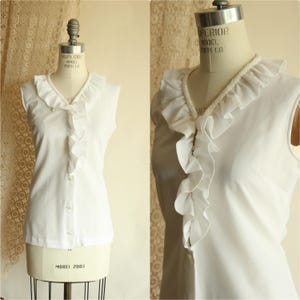 May include: A white sleeveless blouse with a ruffled V-neck and small buttons down the front. The blouse is displayed on a vintage dress form. The fabric looks lightweight and slightly sheer. The style is vintage.