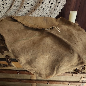 May include: A tan burlap sack featuring a coarse, woven texture and a simple drawstring closure at the top. The fabric is light brown with visible natural fibers, creating a rustic appearance. The sack is folded over at the top opening.