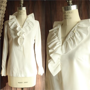 May include: A vintage white blouse with a ruffled collar and cuffs. The long-sleeved shirt is displayed on a mannequin. The blouse has a button closure and a flowing, feminine design, perfect for a retro or classic look.