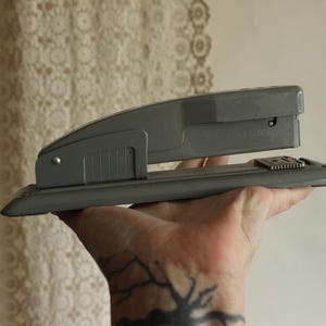 May include: A gray stapler, a common office supply, is held in a hand. The stapler is made of metal and plastic. The background is a cream-colored wall and a lace curtain.