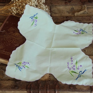 Puede incluir: Un pañuelo bordado en forma de mariposa, verde claro, con bordes festoneados. El pañuelo presenta ramitas bordadas de flores moradas con cintas azules. Un libro vintage es visible en el fondo.