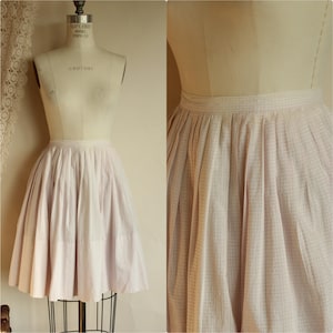 May include: A vintage, light pink skirt with a checkered pattern, displayed on a dress form. The skirt is gathered at the waist, creating a full, flared silhouette. The fabric appears to be a lightweight cotton or blend, perfect for a retro look.
