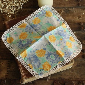 May include: A vintage floral handkerchief with a white lace trim. The handkerchief features a pattern of yellow and blue flowers on a light green background. It is folded and resting on a vintage book.
