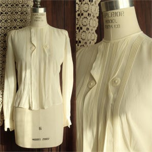 May include: Cream-colored long-sleeve blouse with vertical pleats and decorative buttons. The blouse is displayed on a vintage dress form. The fabric appears to be lightweight, with a high neckline and fitted silhouette. The dress form is labeled "Superior Model Form Co."