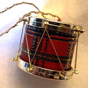May include: A small drum-shaped Christmas ornament with a red tartan pattern and gold detailing. The drum features silver-coloured rims and is suspended by a gold, twisted cord. A festive decoration for the holiday season.