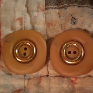 以下が含まれることがあります： 2つの大きくて丸い、タン色のボタン。2つの小さな穴の周りに金色の金属リングが付いています。ボタンは、花柄や模様の生地で作られたパッチワークの背景に配置されています。
