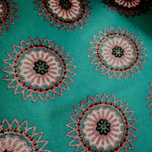 Peut inclure: Gros plan sur un tissu turquoise avec des motifs floraux brodés répétés. Chaque motif circulaire présente un centre blanc avec des accents roses et noirs, entouré d'un contour rose et doré. Le tissu a une texture lisse.