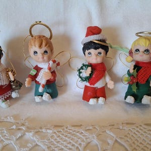 Könnte beinhalten: Vier Weihnachtsengel-Figuren mit Heiligenschein und Flügeln. Jeder Engel trägt ein festliches Outfit, darunter rote, grüne und weiße Kleidung. Ein Engel hält eine Glocke, ein anderer eine Zuckerstange, und zwei weitere halten Kränze und Stechpalmen.