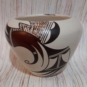 May include: Cream-colored ceramic vase with a wide opening and rounded body. The vase features a brown and black geometric design. The design includes curved lines, shapes, and a textured brown pattern. The vase sits on a light-colored wooden surface.