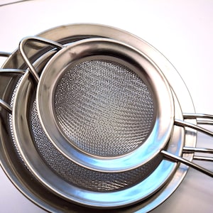 May include: A set of three stainless steel sieves, nested together. Each sieve has a fine mesh screen and a sturdy handle. The sieves are different sizes, with the largest at the bottom and the smallest at the top.