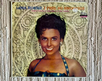 Pode incluir: Uma capa de disco de vinil vintage com Lena Horne, uma famosa cantora e atriz. A capa é de um amarelo vibrante com um design de madeira esculpida no fundo. O texto na capa diz "Lena Horne I Feel So Smoothie" e "Orchestra conducted by Lennie Hayton and Teddy Henderson".