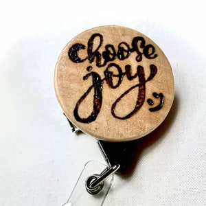 May include: A wooden badge holder with a black retractable cord and a clear plastic strap. The wooden circle has the words "choose joy" written in black.