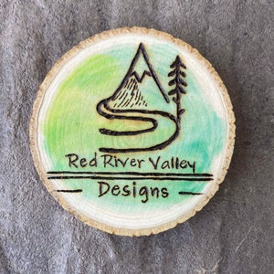May include: A wood slice with a green watercolor background and a black woodburned design of a mountain with a river winding through it. The text "Red River Valley Designs" is also woodburned on the slice.
