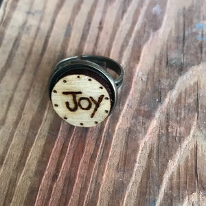 May include: A bronze-toned adjustable ring with a circular wooden face. The face is engraved with the word "Joy" and dotted around the edge. The ring sits on a wooden surface.
