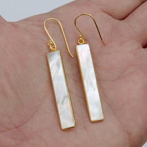 May include: A pair of drop earrings featuring long, rectangular white mother-of-pearl inlays set within thin gold-toned metal frames. Each earring hangs from a simple gold-toned hook wire, showcasing the iridescent, smooth surface of the shell.