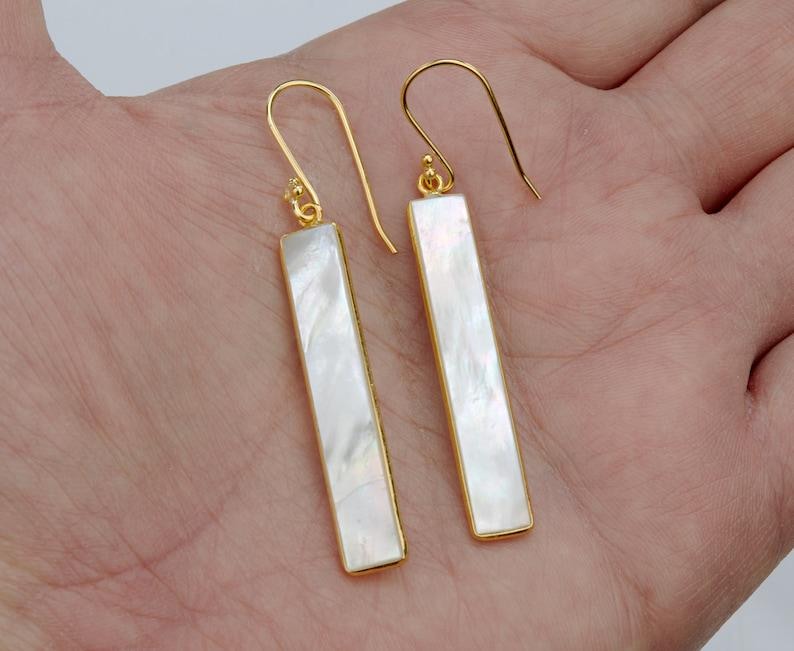 May include: A pair of drop earrings featuring long, rectangular white mother-of-pearl inlays set within thin gold-toned metal frames. Each earring hangs from a simple gold-toned hook wire, showcasing the iridescent, smooth surface of the shell.