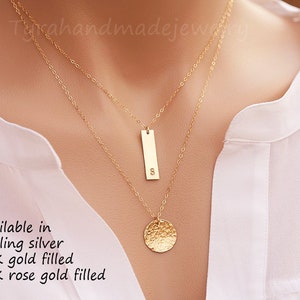 Op de afbeelding: Een gouden ketting met een gehamerde ronde hanger en een staafhanger met de letter 'S' erop. Verkrijgbaar in sterling zilver, 14 karaat goud gevuld en 14 karaat rosé goud gevuld.