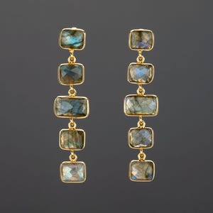 May include: A pair of gold-coloured drop earrings. Each earring has four rectangular, iridescent gemstones in shades of blue, green, and grey. The gemstones are connected by small gold links, creating a dangling effect. The earrings are set against a dark grey background.
