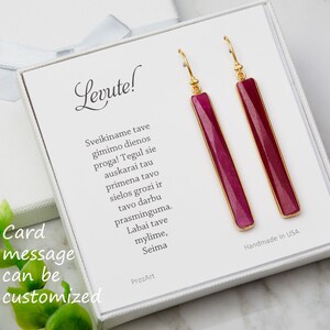May include: A pair of gold earrings with long, rectangular, pink stones. The earrings are in a white box with a message that says "Card message can be customized" and "Levute!" The box also includes a message in Lithuanian.