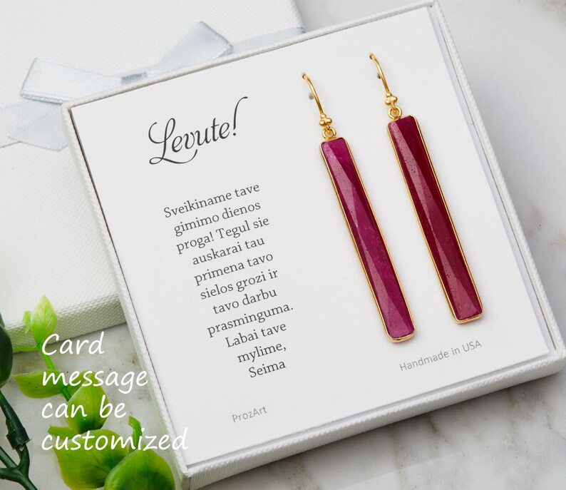 May include: A pair of gold earrings with long, rectangular, pink stones. The earrings are in a white box with a message that says "Card message can be customized" and "Levute!" The box also includes a message in Lithuanian.