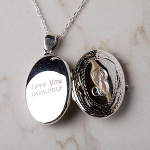 May include: Open silver oval locket pendant on a chain. The locket is engraved with the words "Love You" and the date "12.25.2017" on one side. The other side features an ornate design with the letter "B".