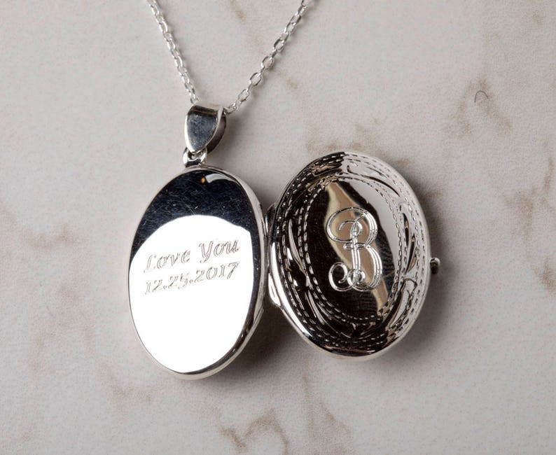 May include: Open silver oval locket pendant on a chain. The locket is engraved with the words "Love You" and the date "12.25.2017" on one side. The other side features an ornate design with the letter "B".