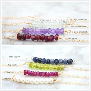 Op de afbeelding: Een set van 12 gouden kettingen met verschillende gekleurde edelsteen kralen. Elke ketting heeft een andere geboortesteen, waaronder granaat, amethist, aquamarijn, witte topaas, saffier, peridoot, robijn en parel. De kettingen worden getoond op een wit oppervlak.