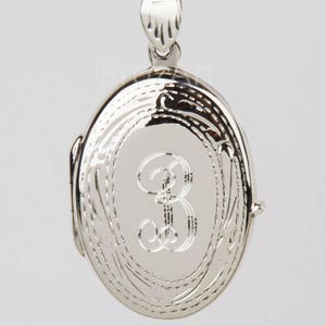 May include: A silver-colored oval locket pendant with a delicate chain. The locket features an engraved letter "B" in the center and decorative etched details around the edges. The pendant is suspended from a fine chain.