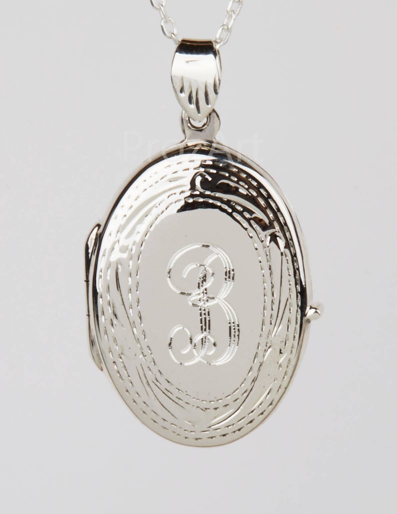 May include: A silver-colored oval locket pendant with a delicate chain. The locket features an engraved letter "B" in the center and decorative etched details around the edges. The pendant is suspended from a fine chain.