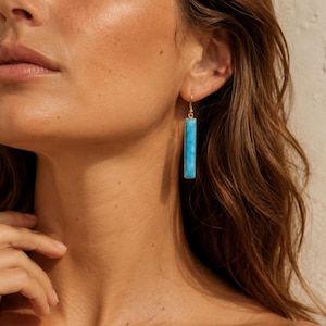 May include: A gold-toned drop earring featuring a long, rectangular, bright turquoise stone pendant. The earring hangs from a thin gold wire hook, showcasing a smooth, polished finish on the vibrant blue stone set within a simple metal frame.