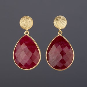 May include: A pair of gold earrings with teardrop-shaped red gemstone accents. The earrings have a textured gold disc at the top.