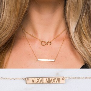 May include: Gold layered necklace with an infinity symbol charm and a bar pendant with the Roman numeral inscription "VI.XVIII.MMXVI".