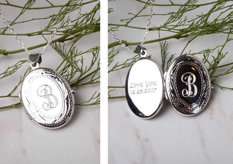 May include: A silver oval locket necklace with an engraved initial "B" and decorative border. The locket opens to reveal the inscription "Love You 12.25.2017". The necklace is displayed against a backdrop of greenery.