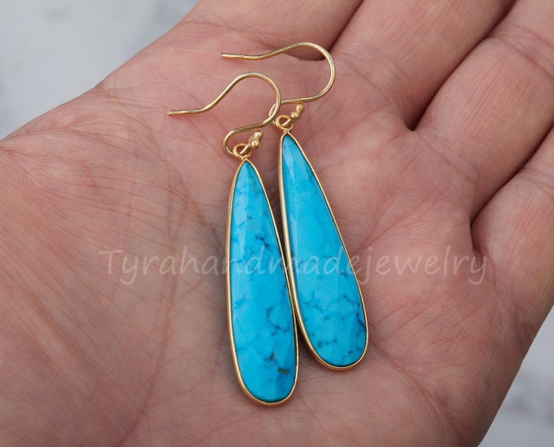 May include: A pair of gold dangle earrings with turquoise teardrop-shaped stones. The earrings have a simple design and are perfect for everyday wear.