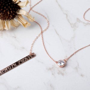 May include: Two rose gold necklaces, one with a small clear gemstone pendant and the other with a bar pendant engraved with the coordinates "41°24'12.2"N 2°10'26.5"E".