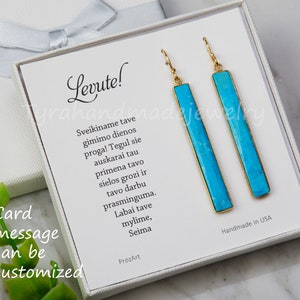 May include: A pair of long, rectangular drop earrings featuring bright turquoise stone inlays set in gold-toned metal frames. The earrings are displayed on a white card with the text Levute and a Lithuanian message, inside a white gift box.