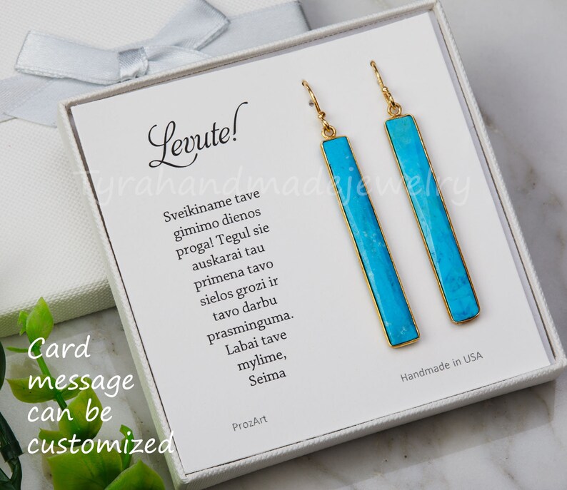 May include: A pair of long, rectangular drop earrings featuring bright turquoise stone inlays set in gold-toned metal frames. The earrings are displayed on a white card with the text Levute and a Lithuanian message, inside a white gift box.