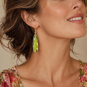 May include: A teardrop-shaped earring featuring a faceted lime green gemstone suspended from a thin gold-toned metal hook. The stone has a pointed bottom and a smooth, reflective surface that catches the light, creating a bright, translucent appearance.
