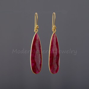 以下が含まれることがあります： 涙型のルビーの宝石が付いた、ゴールドのドロップイヤリングのペア。イヤリングはシンプルなデザインで、毎日の着用に最適です。