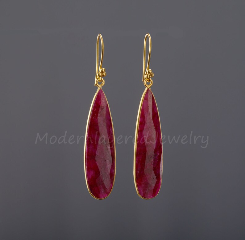 以下が含まれることがあります： 涙型のルビーの宝石が付いた、ゴールドのドロップイヤリングのペア。イヤリングはシンプルなデザインで、毎日の着用に最適です。