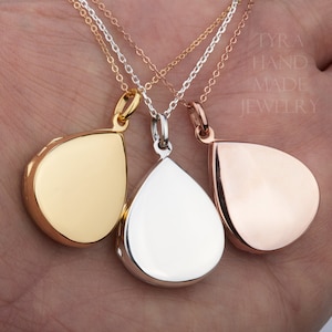 May include: Three teardrop-shaped lockets on chains, one gold, one silver, and one rose gold. The lockets are hanging from a hand.