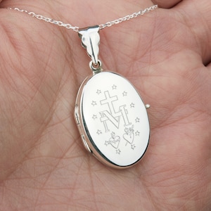 May include: A silver oval locket pendant on a delicate chain. The locket features engraved religious symbols, including a cross, the letters "M" and "N", stars, and two hearts. The pendant is held in a hand.