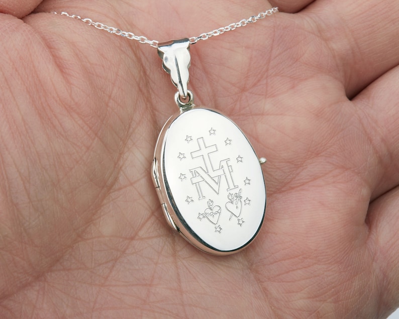 May include: A silver oval locket pendant on a delicate chain. The locket features engraved religious symbols, including a cross, the letters "M" and "N", stars, and two hearts. The pendant is held in a hand.